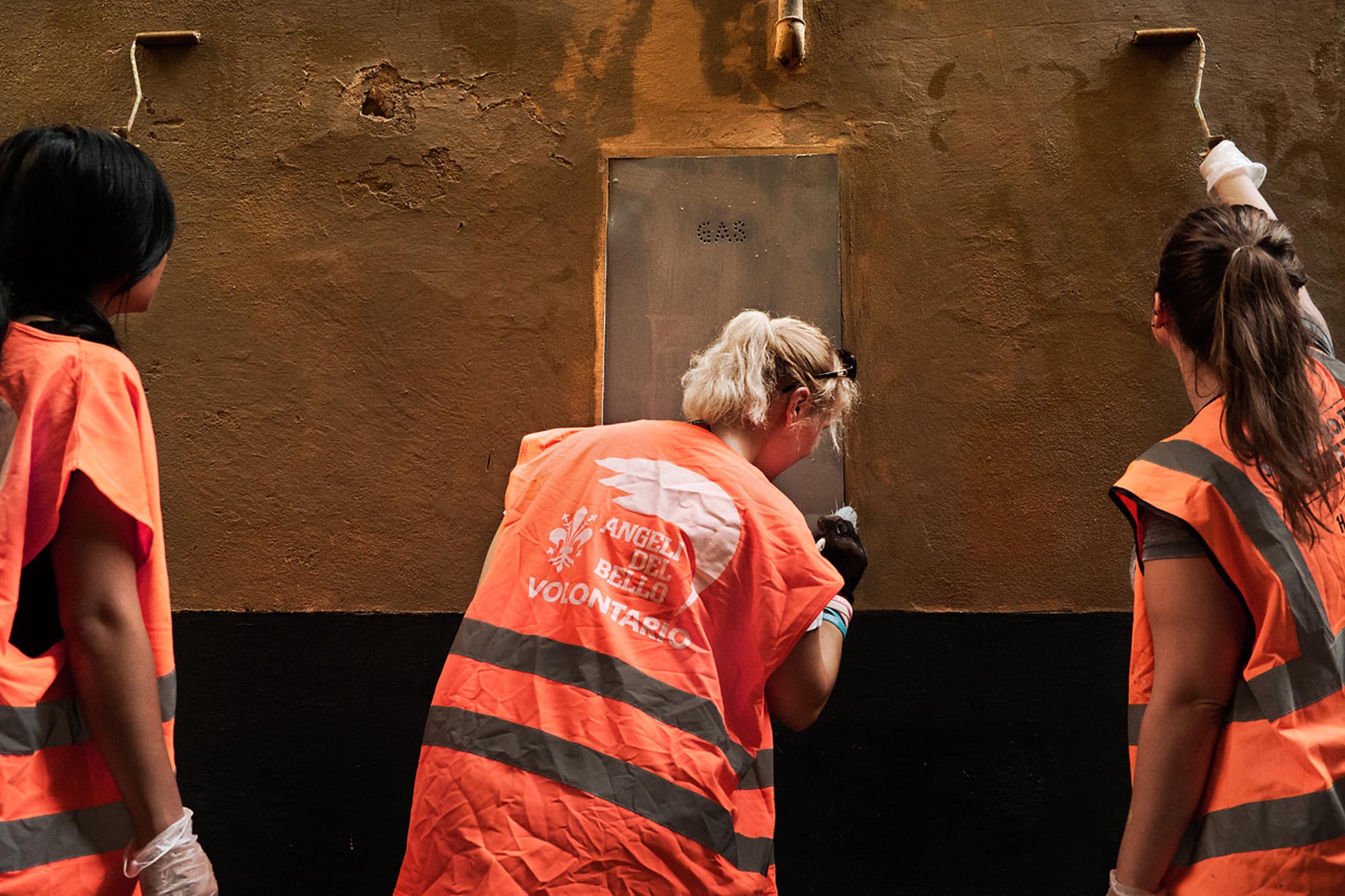 3 persone di spalle con una pettorina fluorescente ripuliscono un muro vandalizzato