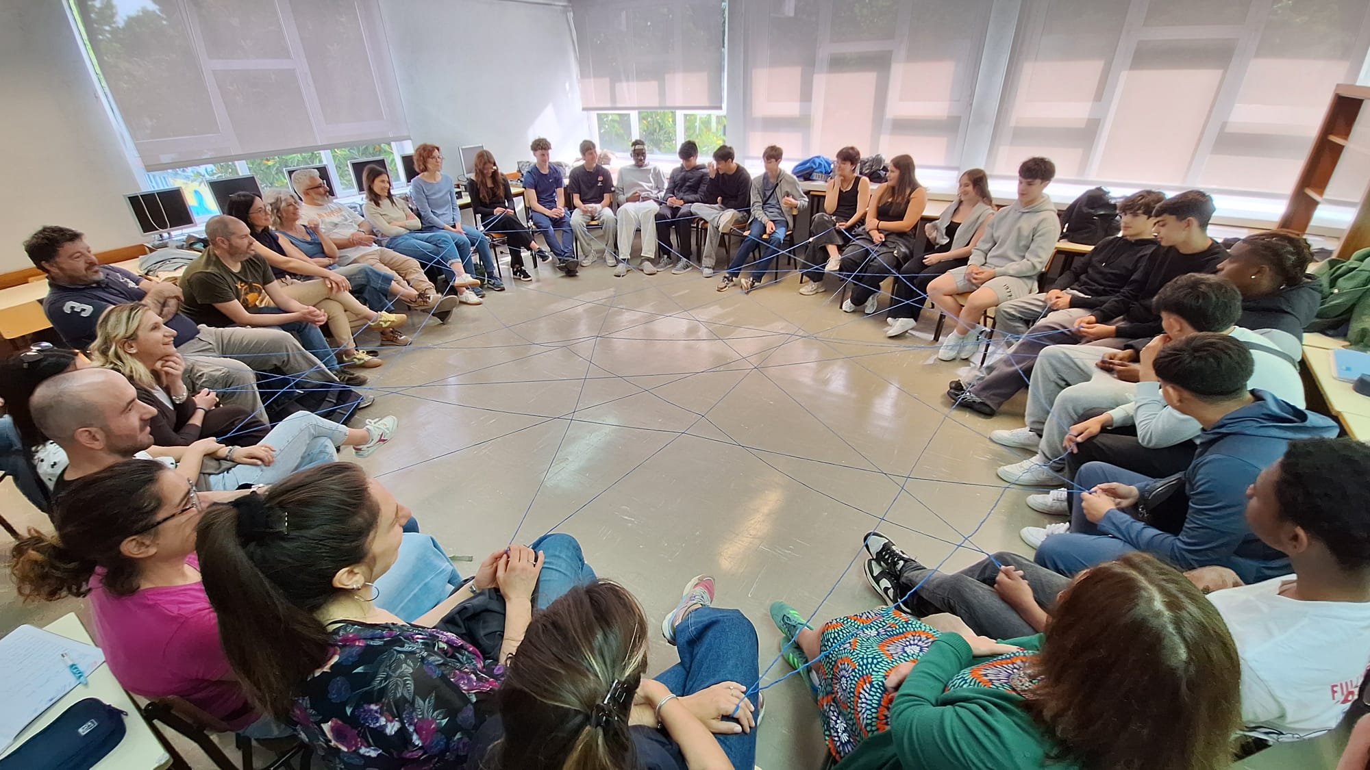 Persone sedute in cerchio, ognuna che tiene il capo di un filo in un intreccio di fili che attravesano il cerchio.