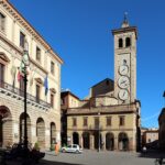 una piazza con torre dell'orologio e palazzo del comune con archi