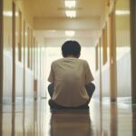 un ragazzino solo seduto a terra di spalle lungo il corridoio di una scuola