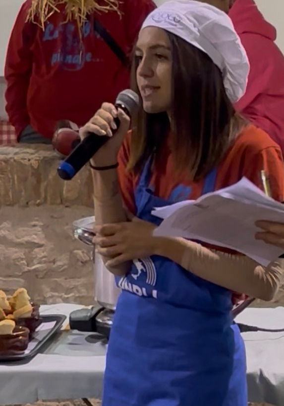 Asya Brescini durante una manifestazione culinaria, con un grembiule addosso, un cappello da cuoco in testa e un microfono in mano