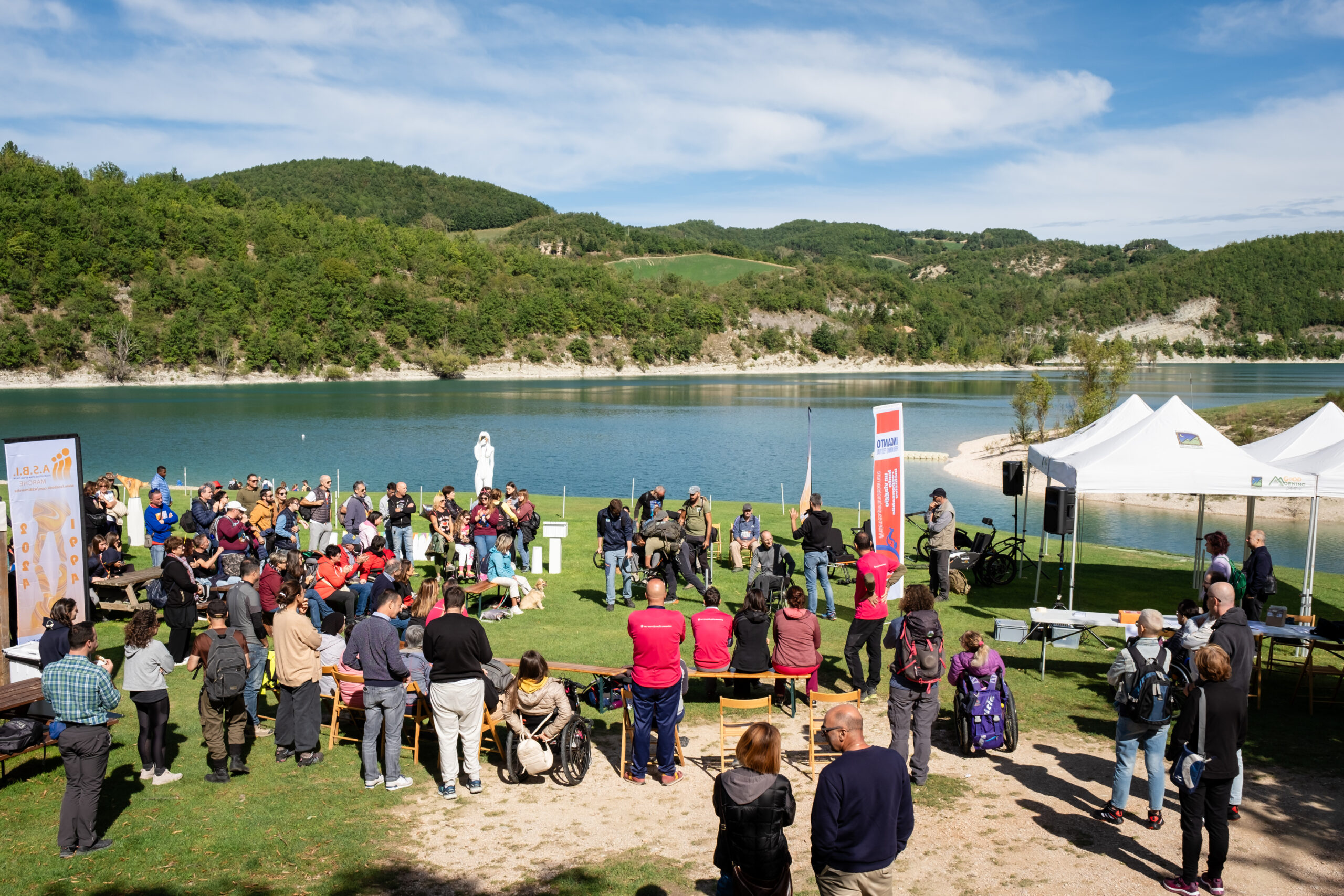 gruppo di persone in un prato a bordo lago partecipa a una presentazione con dimostrazione di joelette