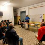 persone sedute in semicerchio ascoltano un relatore in piedi davanti a una grande cartina del territorio