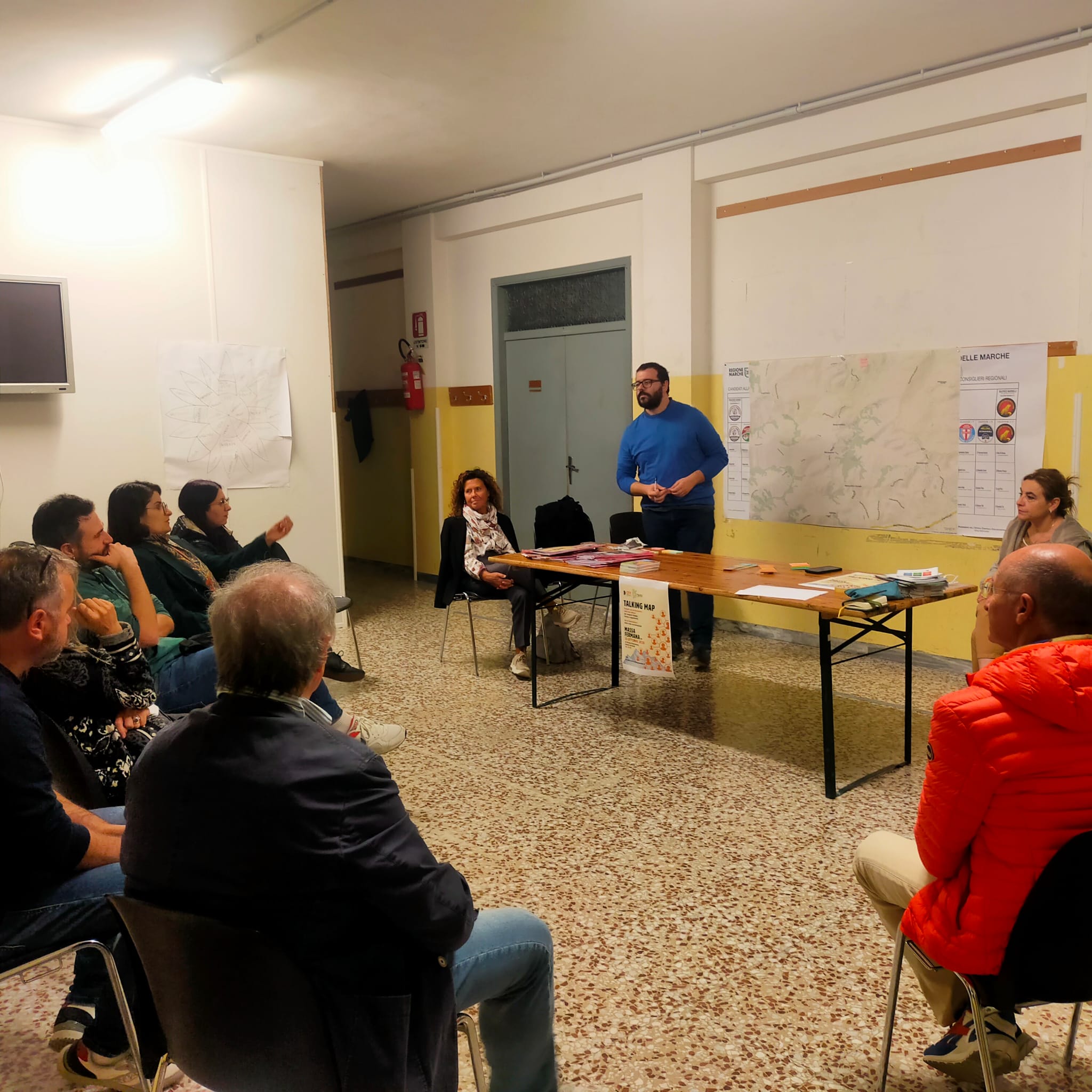 persone sedute in semicerchio ascoltano un relatore in piedi davanti a una grande cartina del territorio