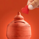 una mano inserisce una moneta a forma di cuore in un salvadanaio rosso