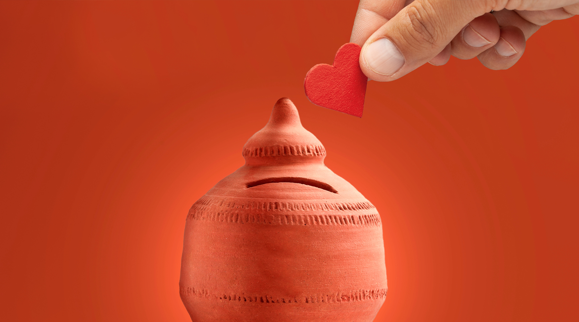 una mano inserisce una moneta a forma di cuore in un salvadanaio rosso