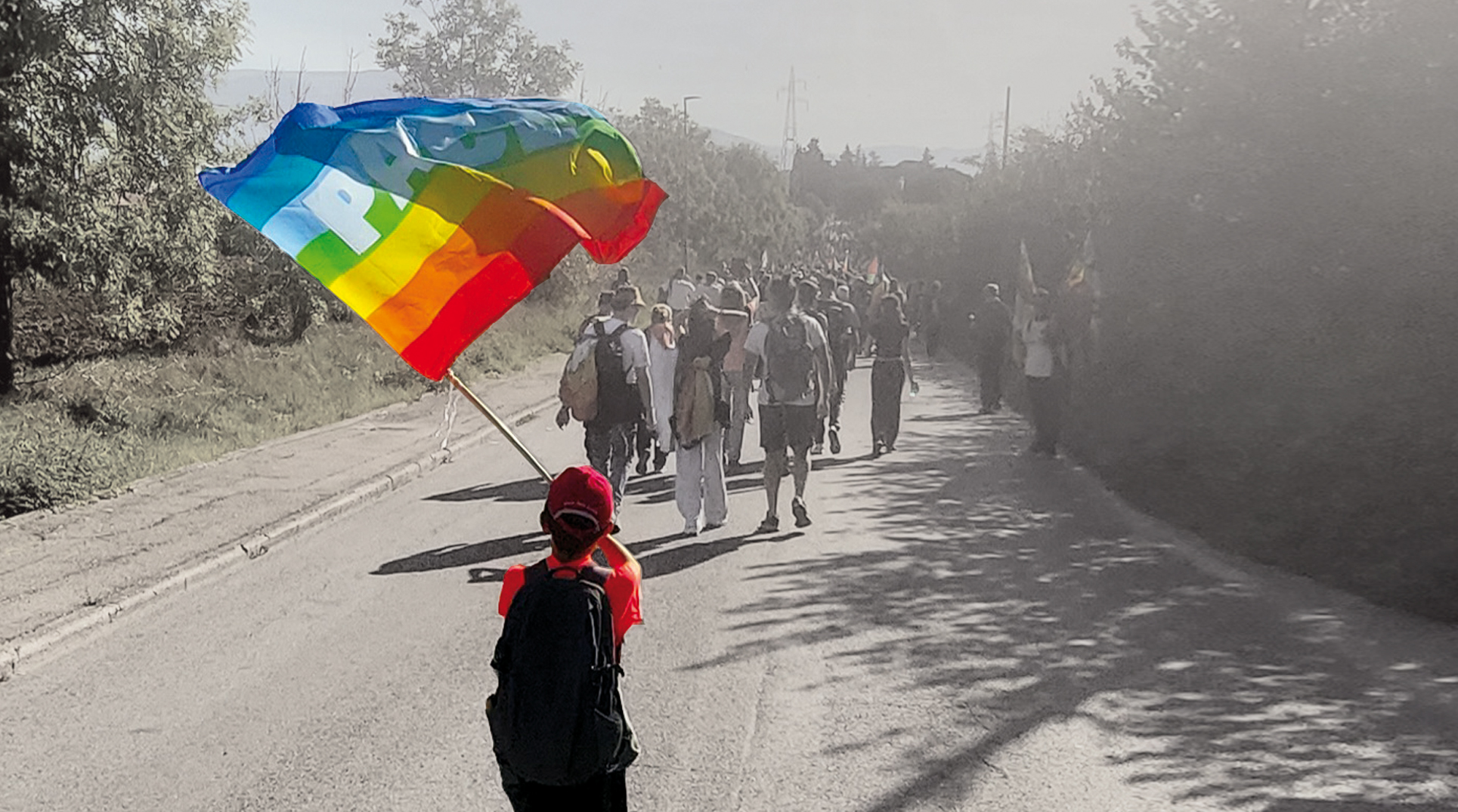 un bambino di spalle sventola la bandiera della pace lungo una strada. La sua figura è a colori, tutto il resto è in bianco e nero.