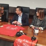 i 3 relatori della conf stampa dietro il tavolo, con appoggiate le t shirt colorate e altri gadget del progetto