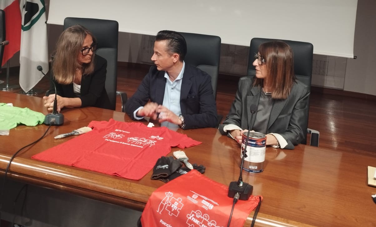 i 3 relatori della conf stampa dietro il tavolo, con appoggiate le t shirt colorate e altri gadget del progetto