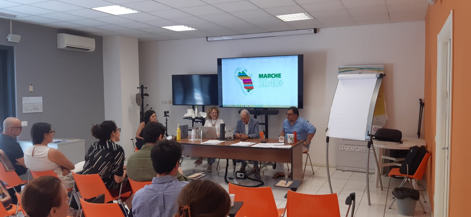 una sala con persone in platea e quattro relatori sotto un grande monitor